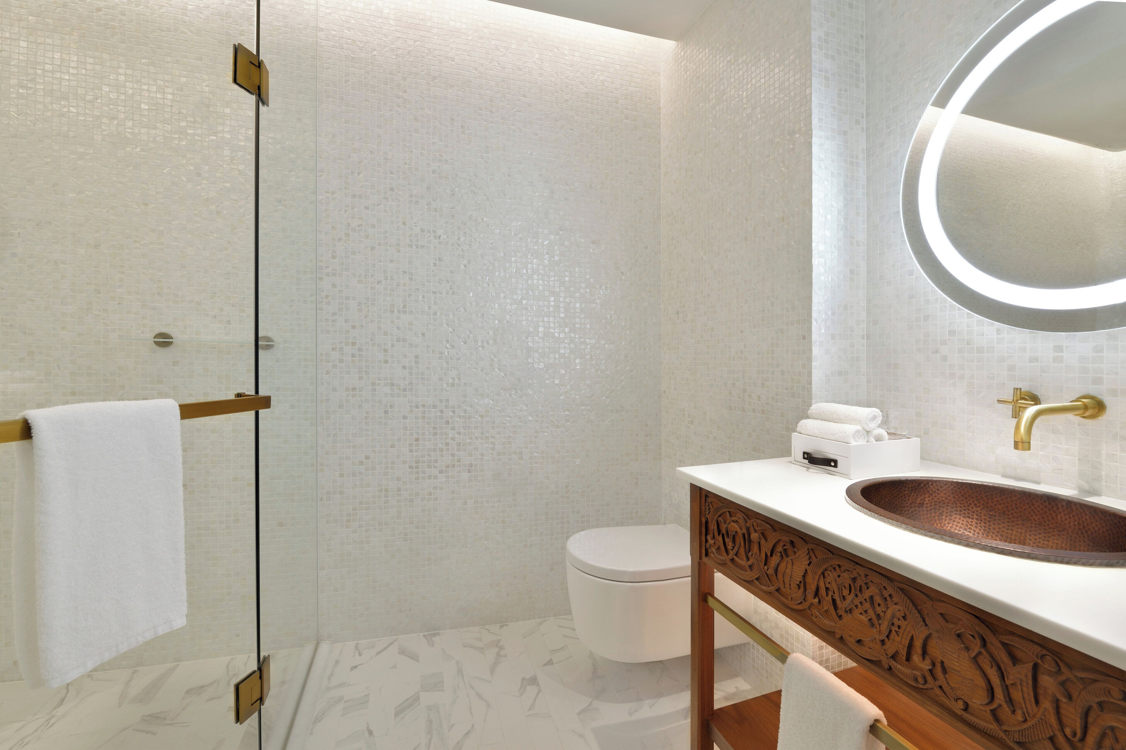 A bathroom with a glass shower enclosure, a white toilet, a wooden vanity with a copper sink, and a round mirror.