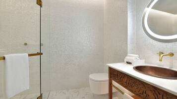 A bathroom with a glass shower enclosure, a white toilet, a wooden vanity with a copper sink, and a round mirror.