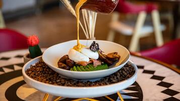 A bowl of food with a sauce being poured over it, placed on a table with a patterned tablecloth.