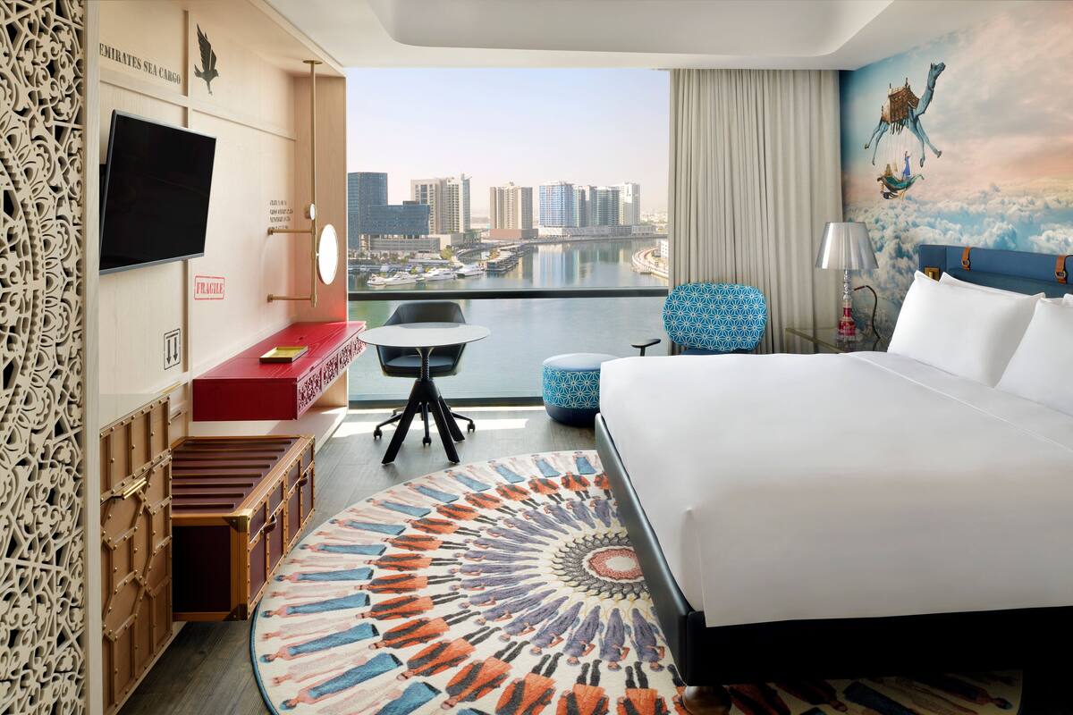 Ein Hotelzimmer mit einem großen Bett, einem Schreibtisch mit Stuhl, einem kleinen Tisch, einem an der Wand befestigten Fernseher und Blick auf die Stadt.