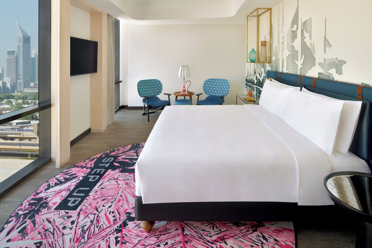 Ein Hotelzimmer mit einem großen Bett, zwei Stühlen, einem kleinen Tisch mit einer Lampe und Blick auf die Stadt.