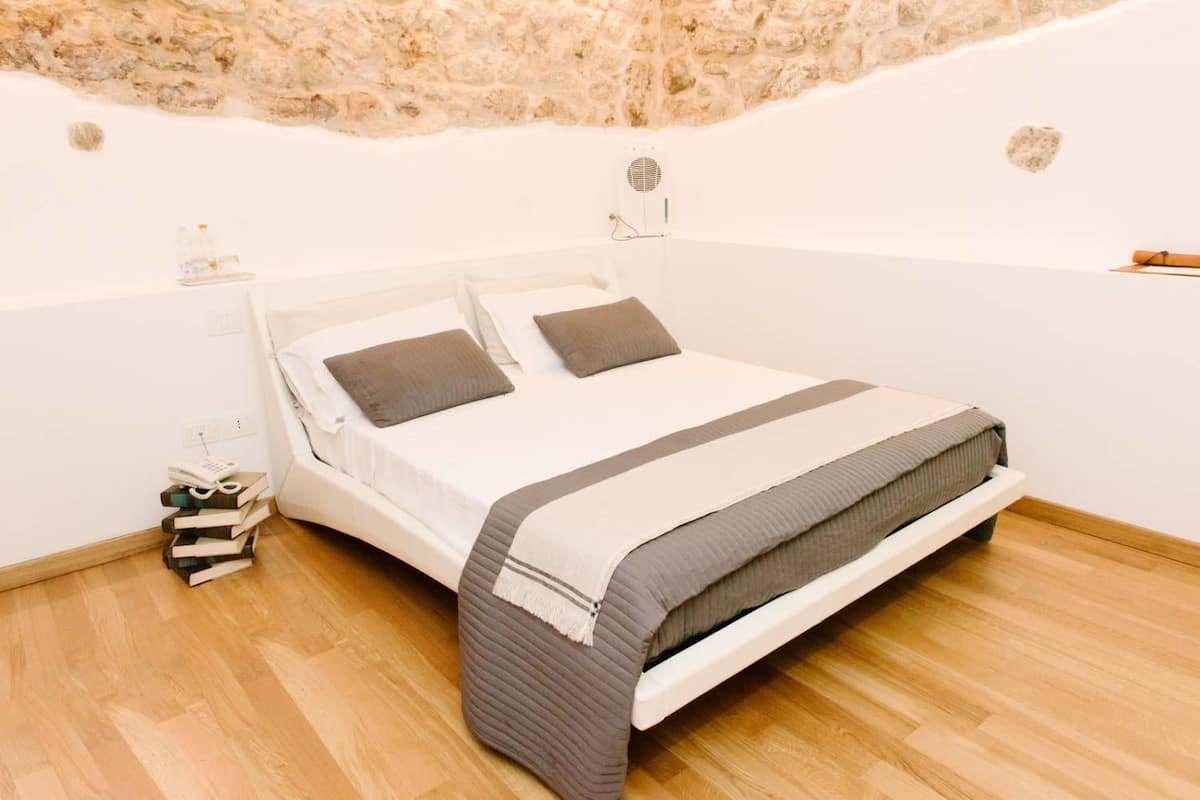 A single bed with a white frame and grey bedding, a small bedside table with a lamp, and a stack of books on the floor.