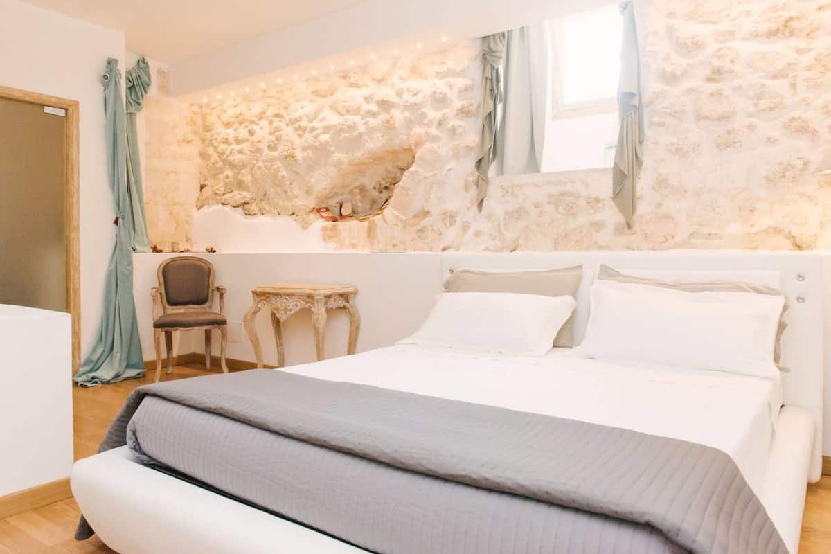 A bedroom with a stone wall, a bed with white bedding, a small table, and a chair.