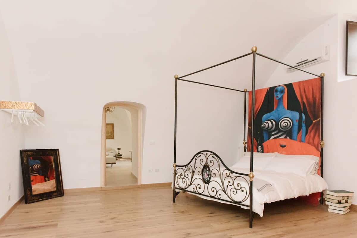 A four-poster bed with a white bedspread and red pillows, a framed picture on the wall, and a large abstract painting above the bed.