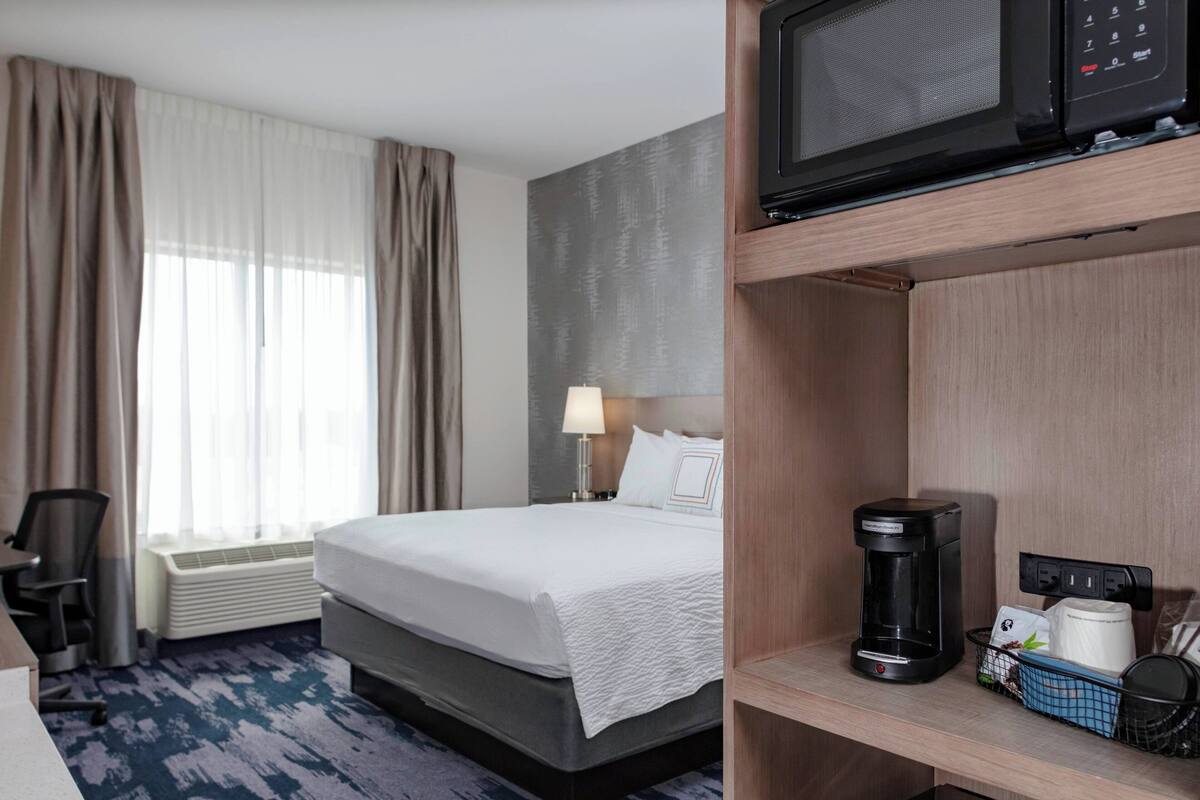 Ein Hotelzimmer mit Bett, Schreibtisch, Stuhl, Kaffeemaschine, Mikrowelle und einem fenster mit Vorhängen.