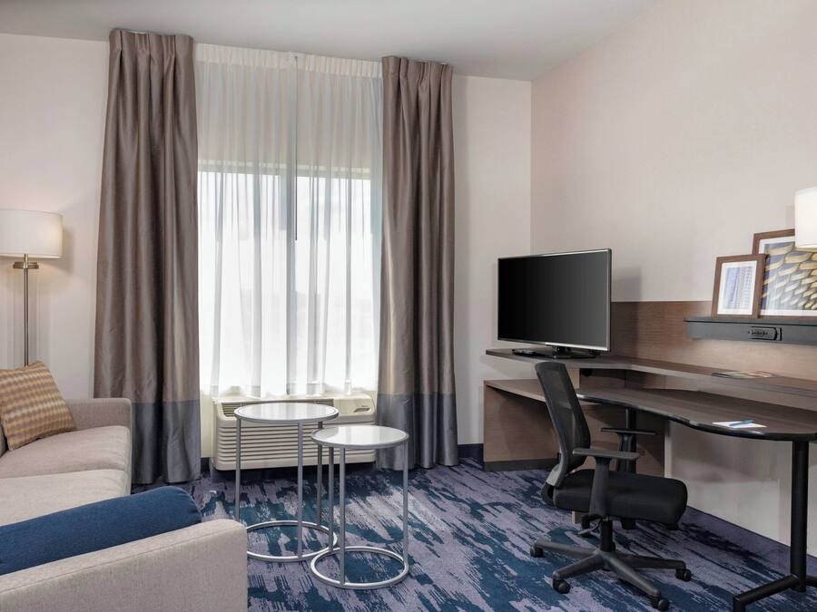 Ein Hotelzimmer mit einer Couch, einem Schreibtisch mit Computer, einem Fernseher, einem kleinen Tisch und einem Fenster mit Vorhängen.