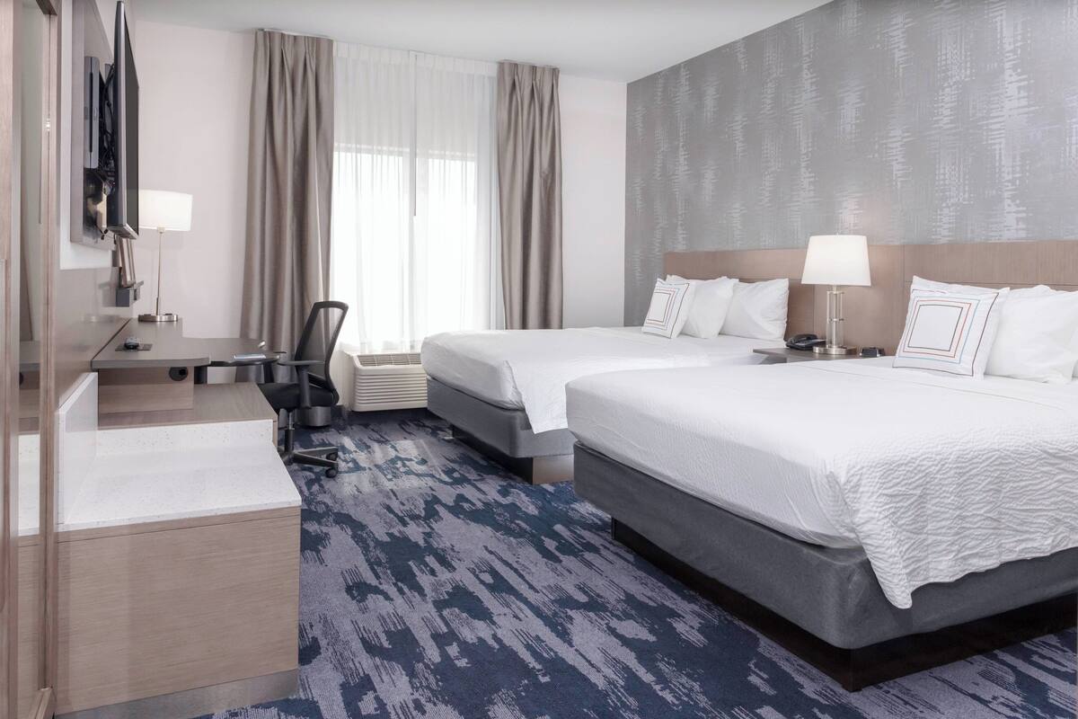 Ein Hotelzimmer mit zwei Betten, einem Schreibtisch, einem Stuhl, einem Fernseher und einem großen Fenster mit Vorhängen.