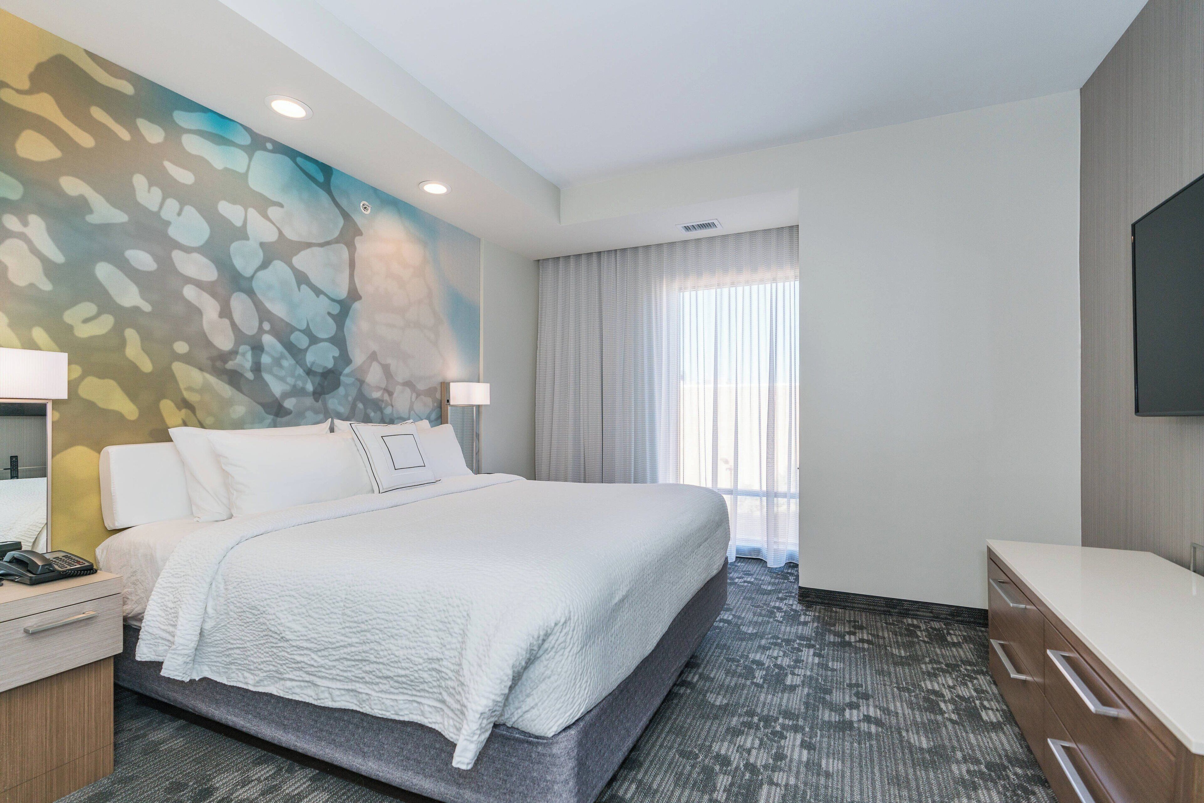 A modern hotel room with a large bed, a desk with a telephone, a flat-screen TV, and a window with curtains.