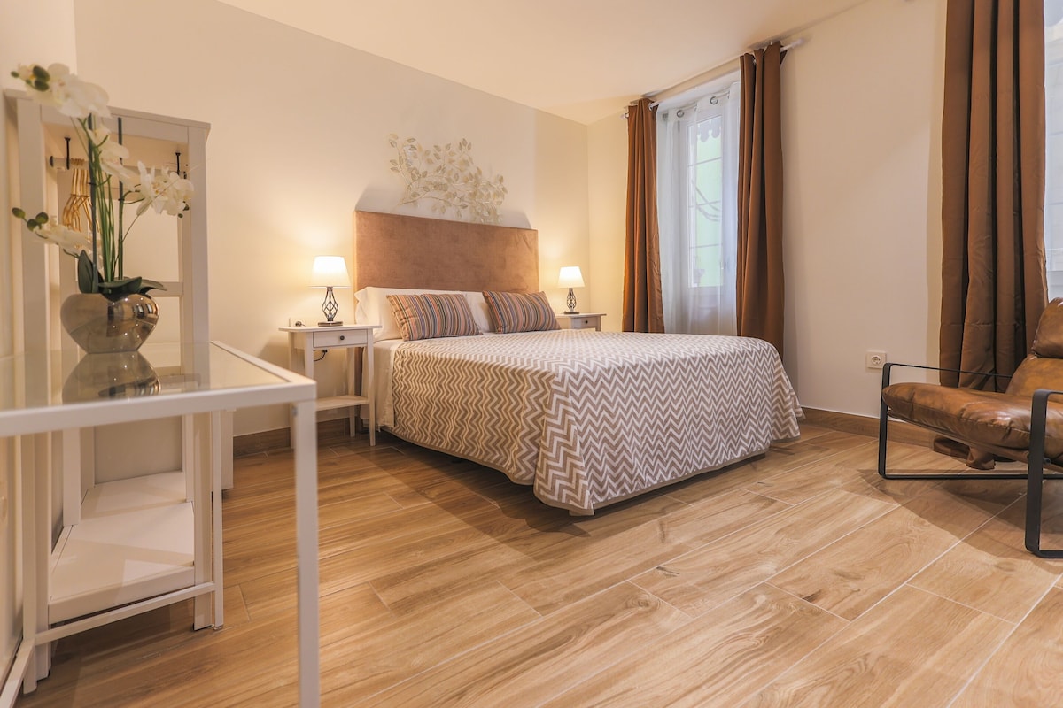 A modern bedroom with a large bed, a bedside table with lamps, a chair, and a decorative wall piece.