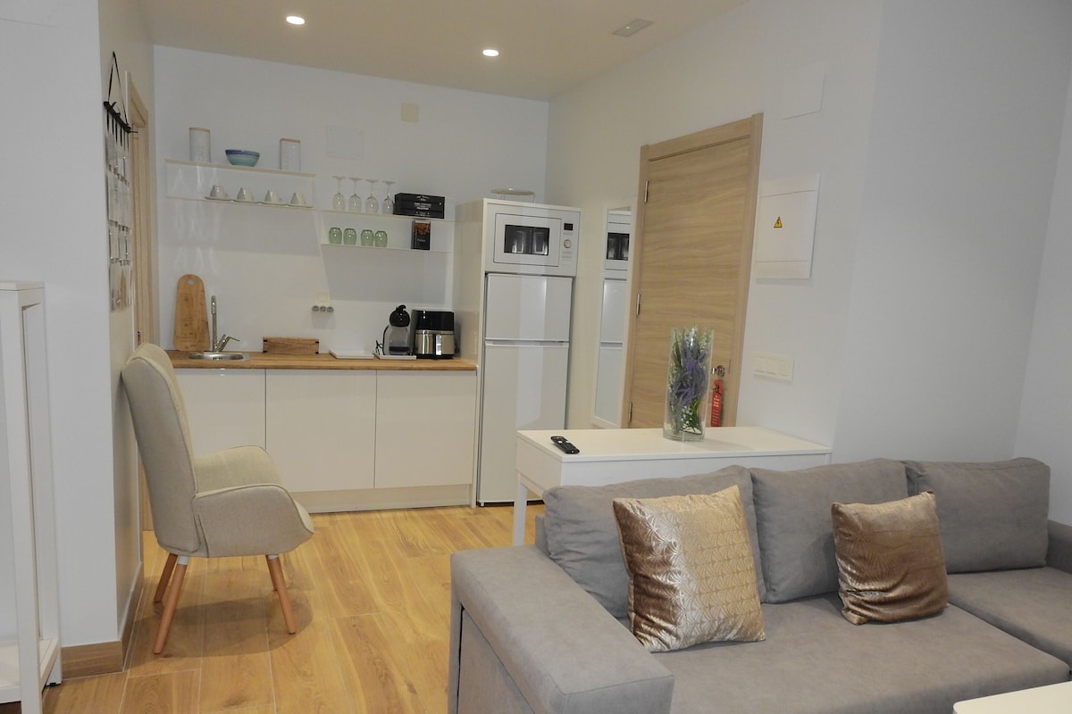 A modern kitchen with white cabinets, a coffee maker, and a refrigerator. There is a grey sofa with two cushions and a wooden chair.