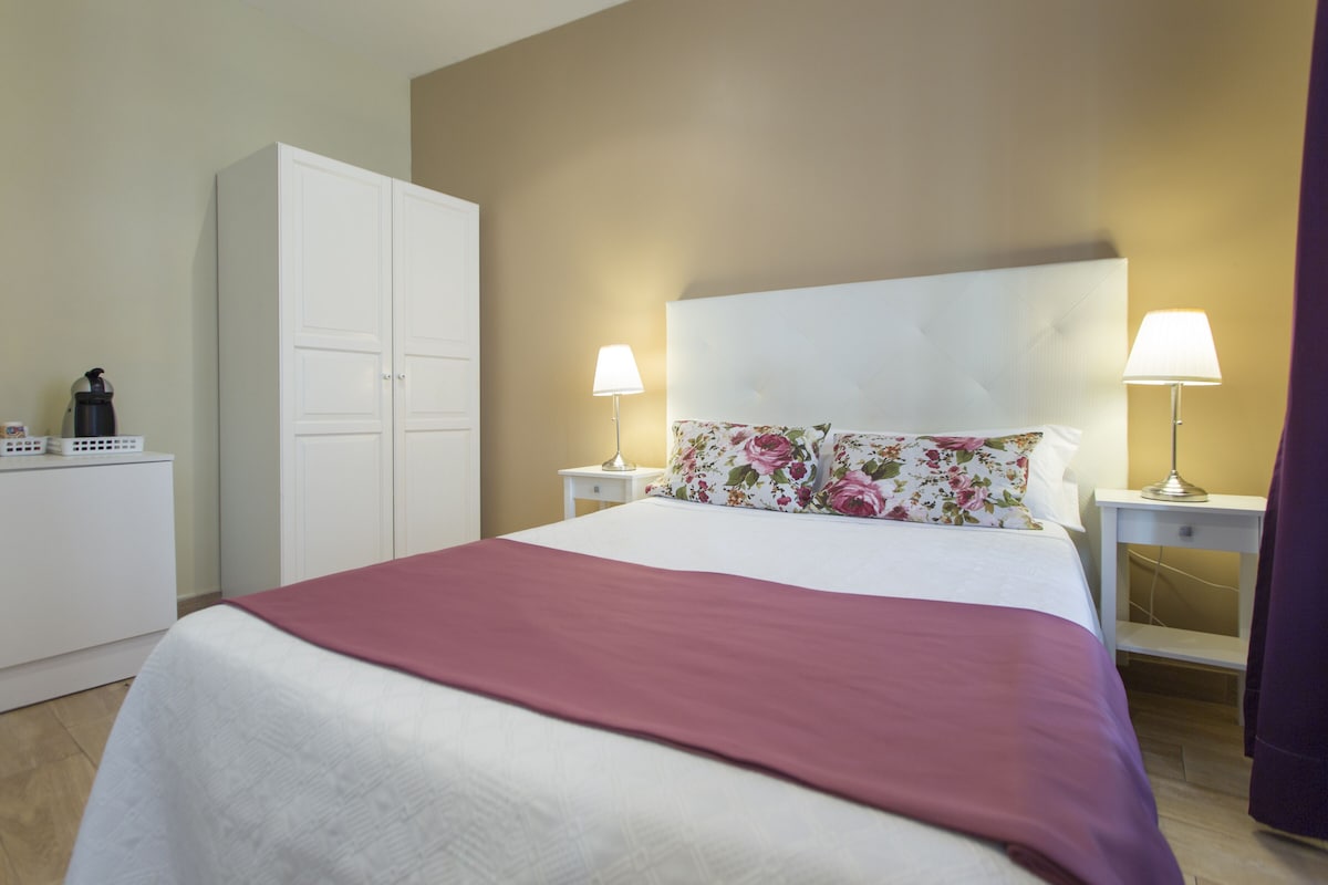 A neatly made bed with a white and maroon bedspread, two bedside lamps, a floral pillow, and a white wardrobe.