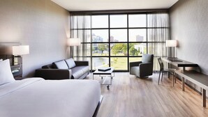 A modern hotel room with a large bed, a sofa, a desk, and a view of a cityscape through large windows. - AC Hotel by Marriott Huntsville Downtown (Huntsville)