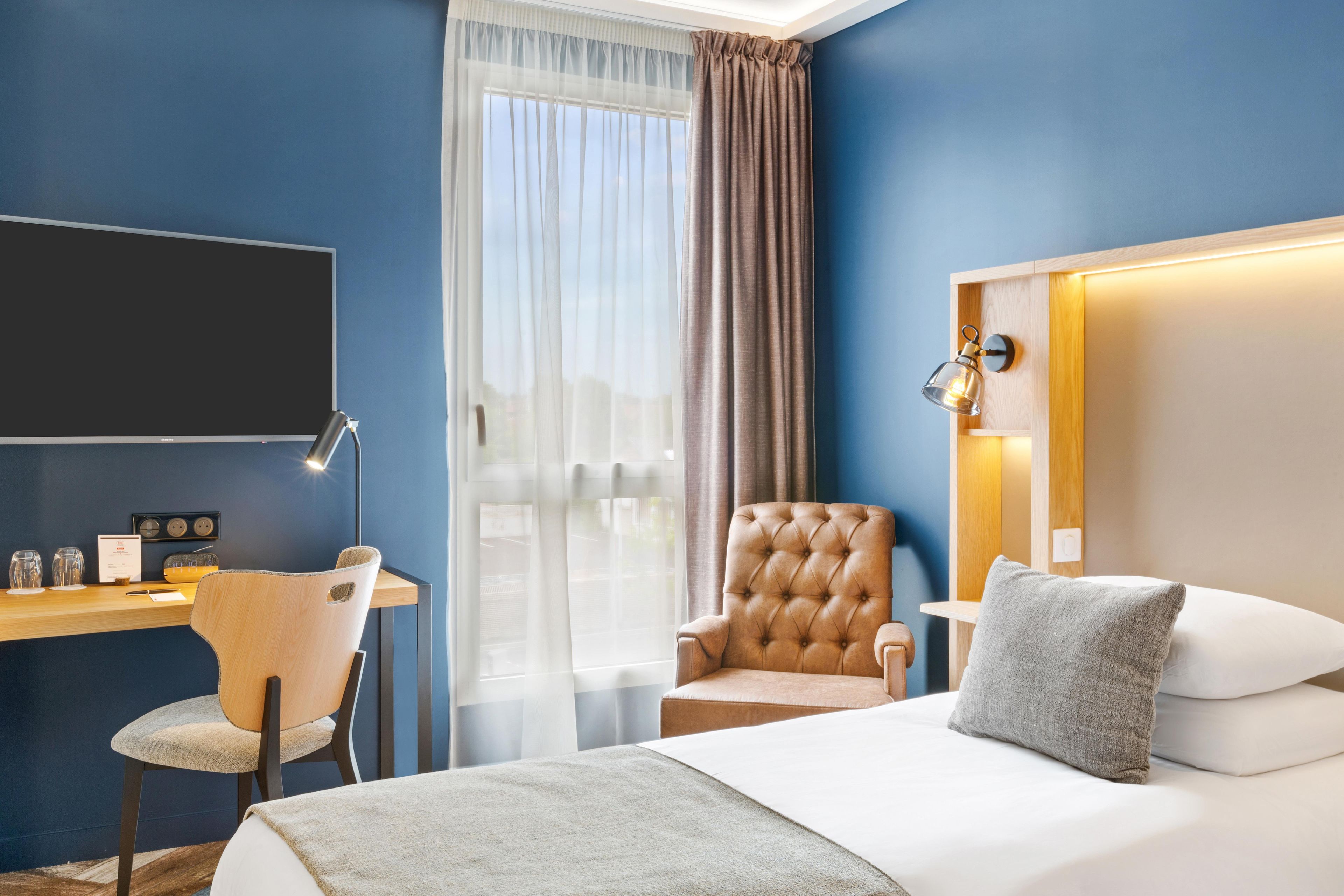 Quarto de hotel com cama, escrivaninha e cadeira, televisão e janela com cortinas.