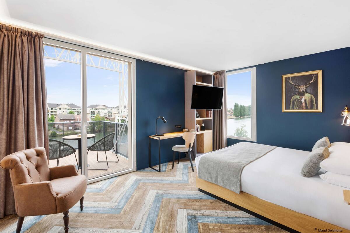 Une chambre à coucher comprenant un lit, un bureau, une chaise, un téléviseur et une grande fenêtre donnant sur des immeubles.