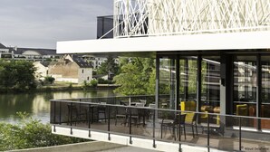 A modern building with a glass balcony overlooking a river, featuring outdoor seating and a view of the surrounding area.