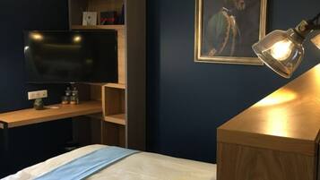 A bedroom with a bed, a desk with a computer, a television, a shelf with items, and a framed picture on the wall.