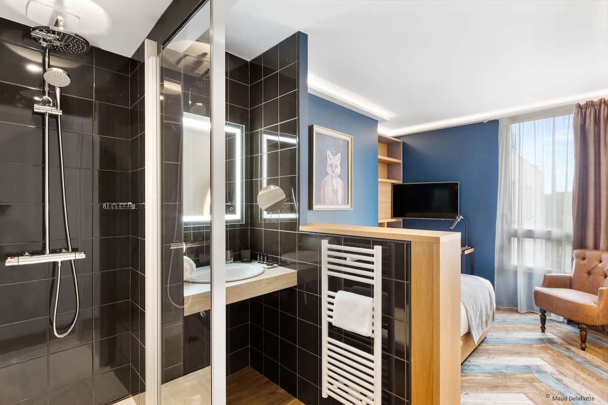 Une salle de bain moderne dans un hôtel, dotée d’une douche en verre, d’un lavabo et d’un meuble-lavabo. La pièce adjacente comprend un lit, une télévision et une chaise.