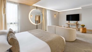 A modern hotel room with a large bed, a round mirror, a flat-screen TV, and a seating area with a wooden side table.