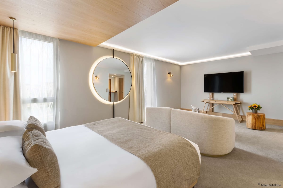 Une chambre d’hôtel moderne dotée d’un grand lit, d’un miroir rond, d’un téléviseur à écran plat et d’un coin salon avec une table d’appoint en bois.