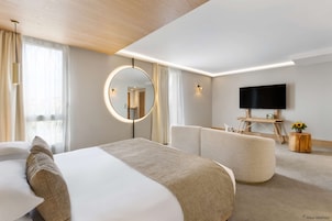 Quarto de hotel moderno com uma cama grande, um espelho redondo, uma TV de tela plana e uma área de estar com uma mesinha de madeira.