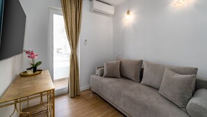 A living room with a beige sofa, a wooden coffee table, and a small potted plant. There is a window with curtains, an air conditioning unit, and wall-mounted lighting. - Rooms Vedrana (Podstrana)