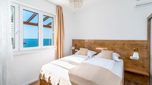 A bedroom with a large bed, wooden headboard, a view of the sea through a window, and a chandelier. - Rooms Vedrana (Podstrana)