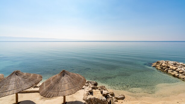 A beach with clear water, a rocky shoreline, and straw umbrellas. - Rooms Vedrana (Podstrana)