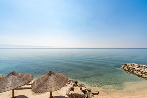 A beach with clear water, a rocky shoreline, and straw umbrellas. - Rooms Vedrana (Podstrana)
