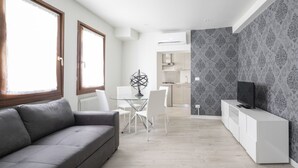 A modern living room with a grey sofa, a dining table with chairs, a kitchen area with a TV on a white cabinet, and patterned wallpaper. - Apartments in San Marco with Canal View by Wonderful Italy (Venice)