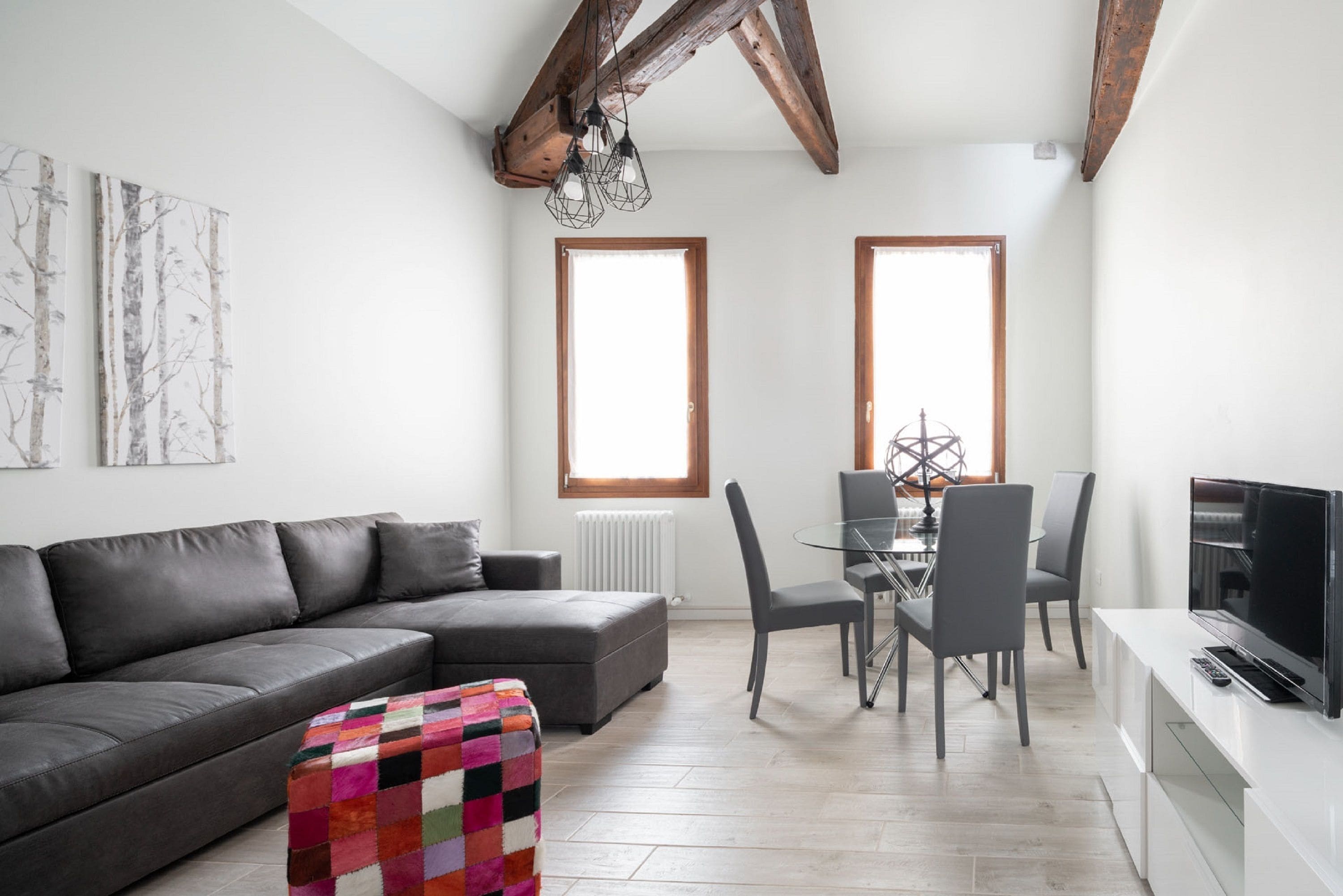 Una sala moderna con un sofá gris, un otomano colorido, un comedor con una mesa de vidrio y sillas, y un televisor sobre un mueble blanco.