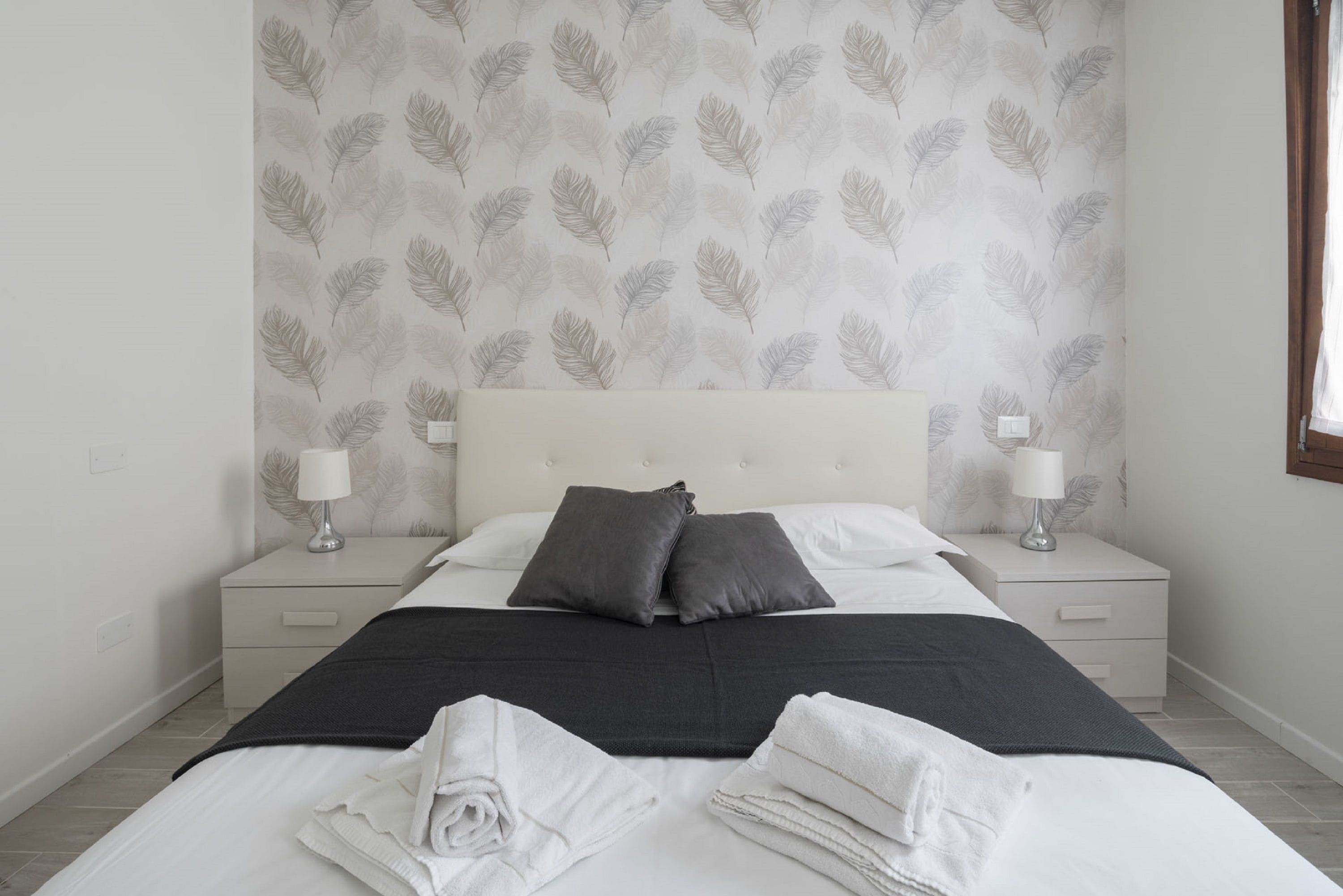Een net opgemaakt bed met kussens en een deken, twee nachtkastjes met lampen, en een muur met een bladmotief.