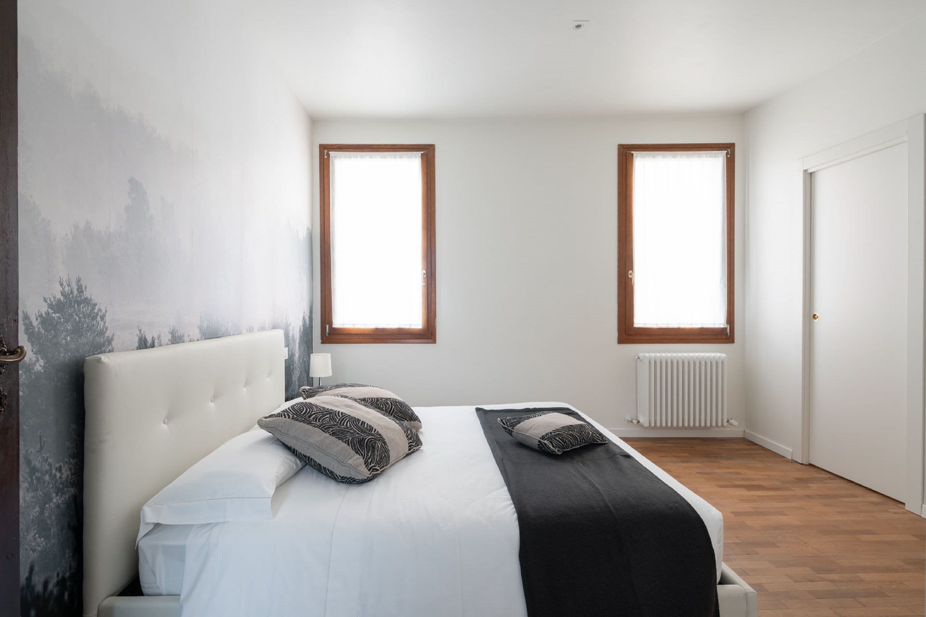 A bedroom with a large bed, two windows, a radiator, and a door.