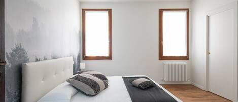 Ein Schlafzimmer mit einem großen Bett, zwei Fenstern, einer Heizung und einer Tür.