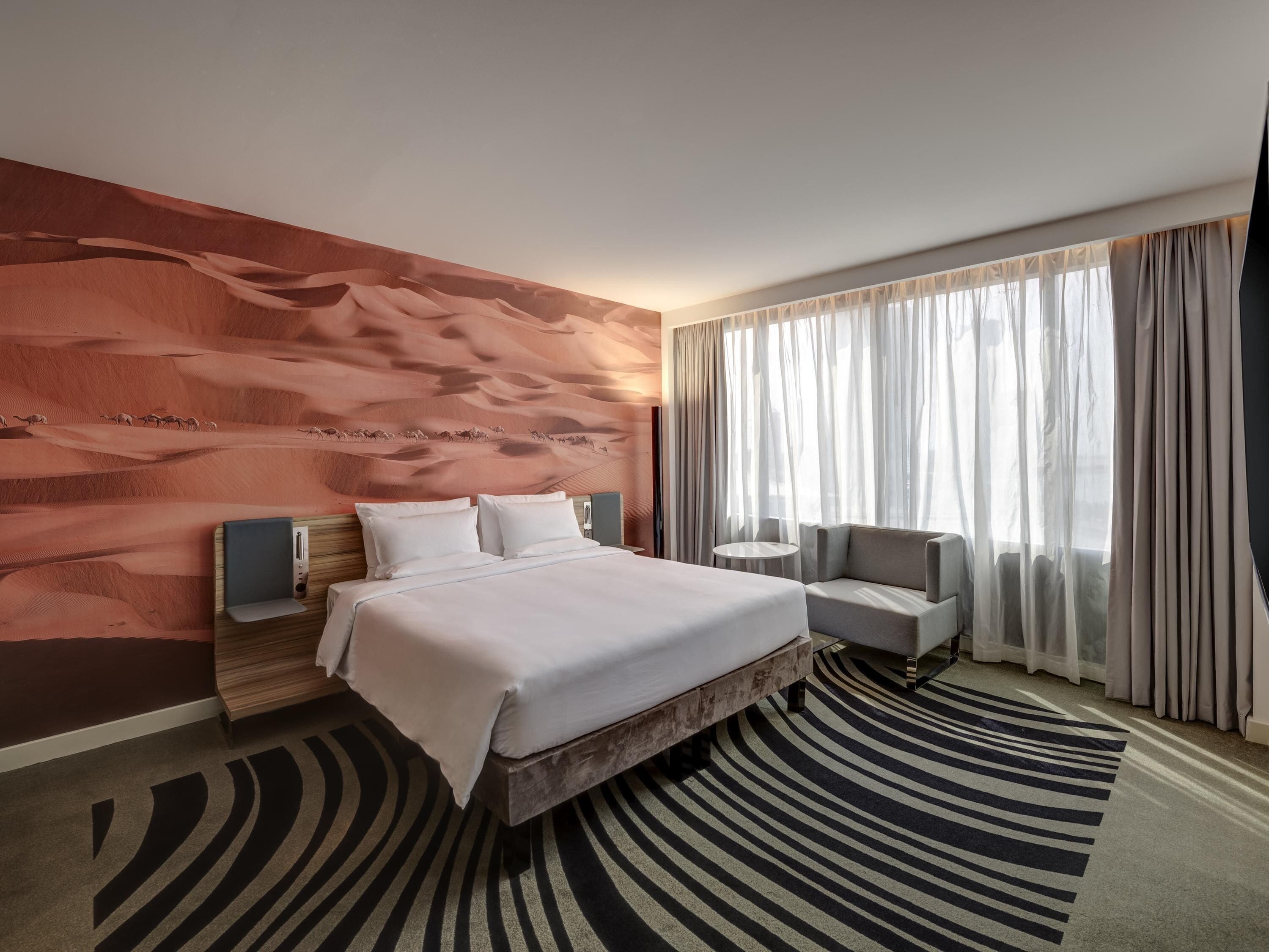 Ein Hotelzimmer mit einem Bett, einem Sessel, einem runden Tisch, einem großen Fenster mit Vorhängen und einem Wandgemälde.
