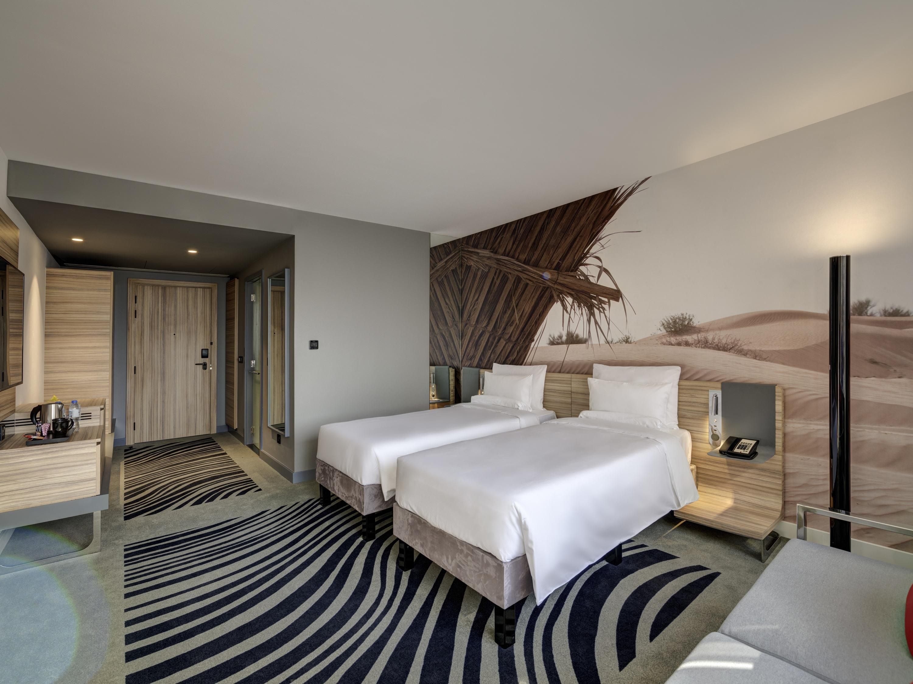 A hotel room with two beds, a desk with a chair, a small table, a nightstand, and a wall mural.