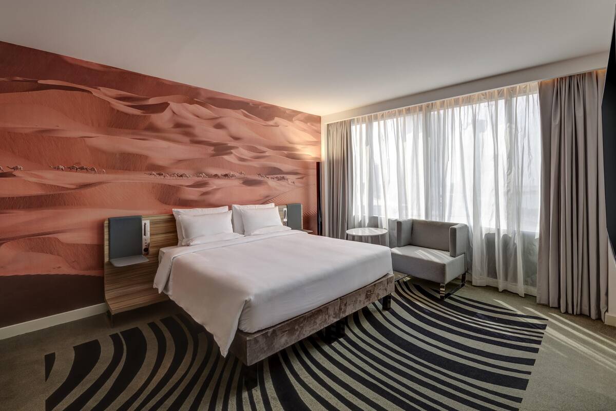 Habitación de hotel con cama, una silla, una mesa redonda, un ventanal con cortinas y un mural en la pared.