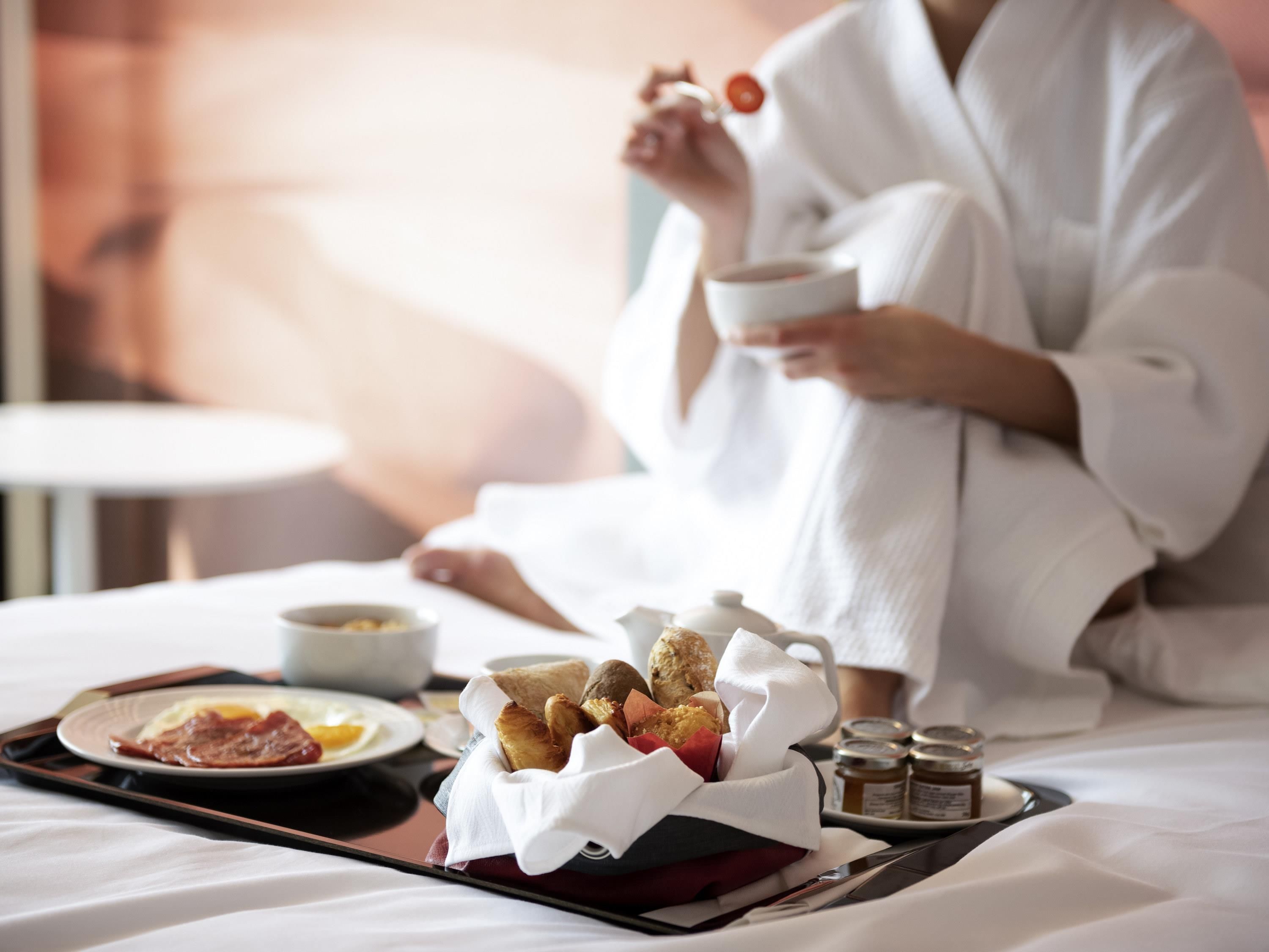 A person in a white robe sitting on a bed with breakfast items including toast, eggs, and a cup.