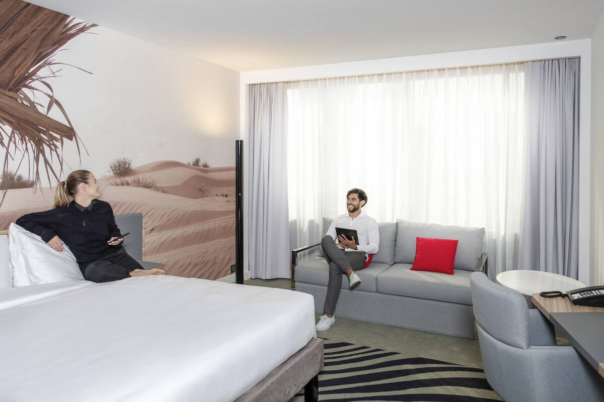 A hotel room with a bed, a sofa, a small table, and a large window with curtains.
