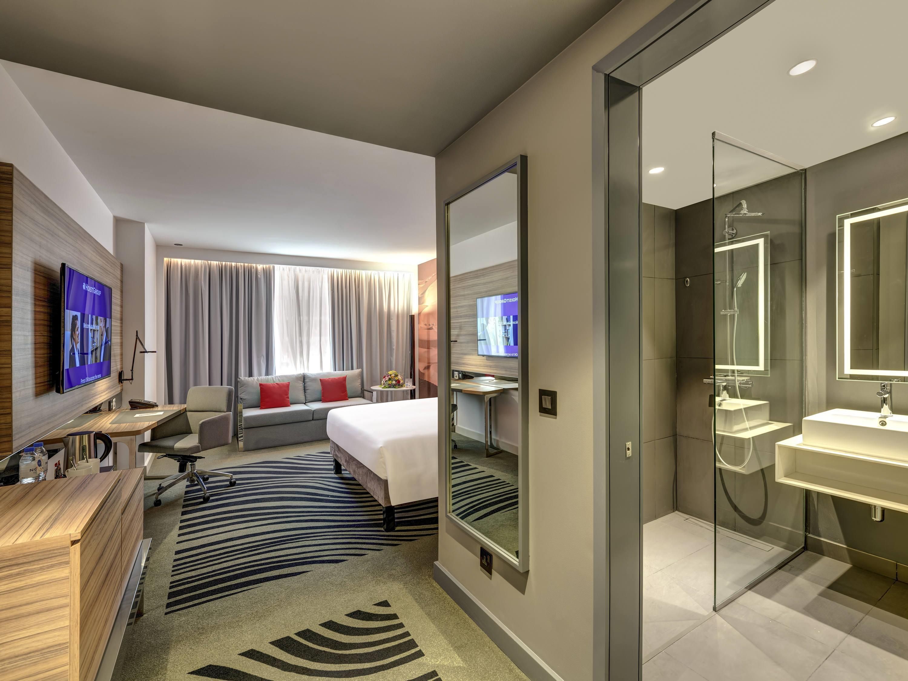 Ein modernes Hotelzimmer mit einem großen Bett, einem Schreibtisch mit Computer, einem Fernseher, einer Couch und einem Badezimmer mit Glasdusche.