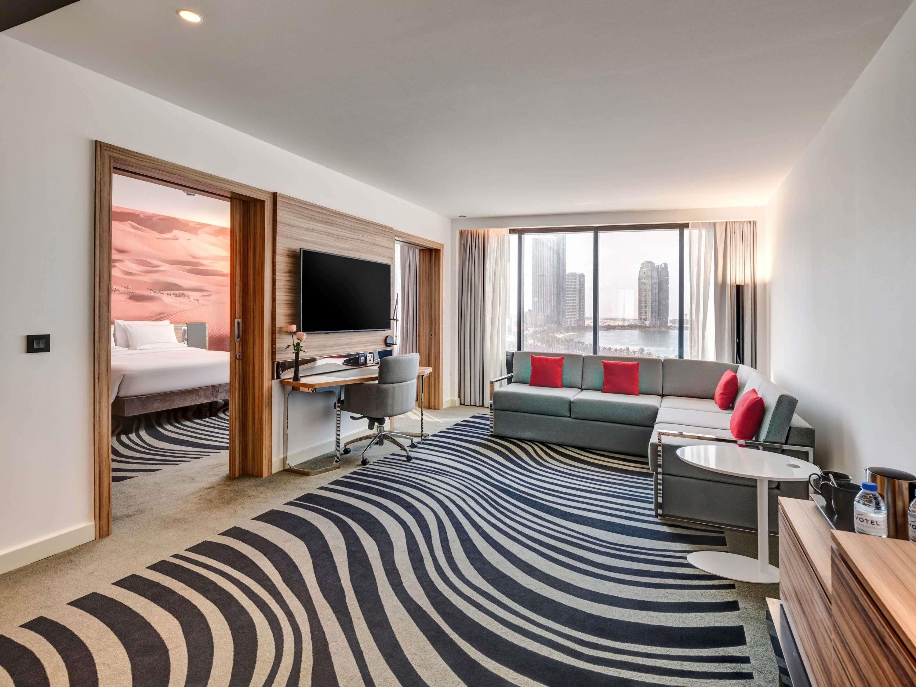 A hotel room with a bed, desk, TV, sofa, and a view of the city.