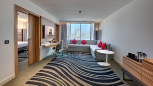 A modern hotel room with a large window, a sofa with red cushions, a desk with a chair, a small round table, and a wooden cabinet. - Novotel Sharjah Expo Center (Sharjah)
