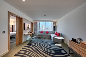 Una habitación de hotel moderna con un ventanal grande, un sofá con cojines rojos, un escritorio con una silla, una mesita redonda pequeña y un armario de madera.