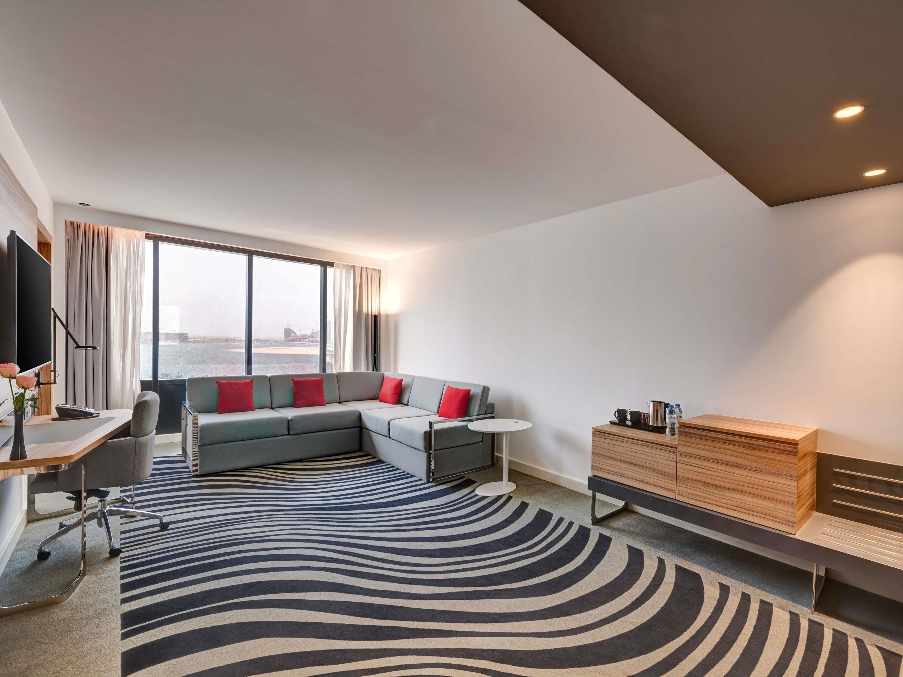 A modern hotel room with a large window, a sofa, a desk, a small table, and a wooden cabinet.