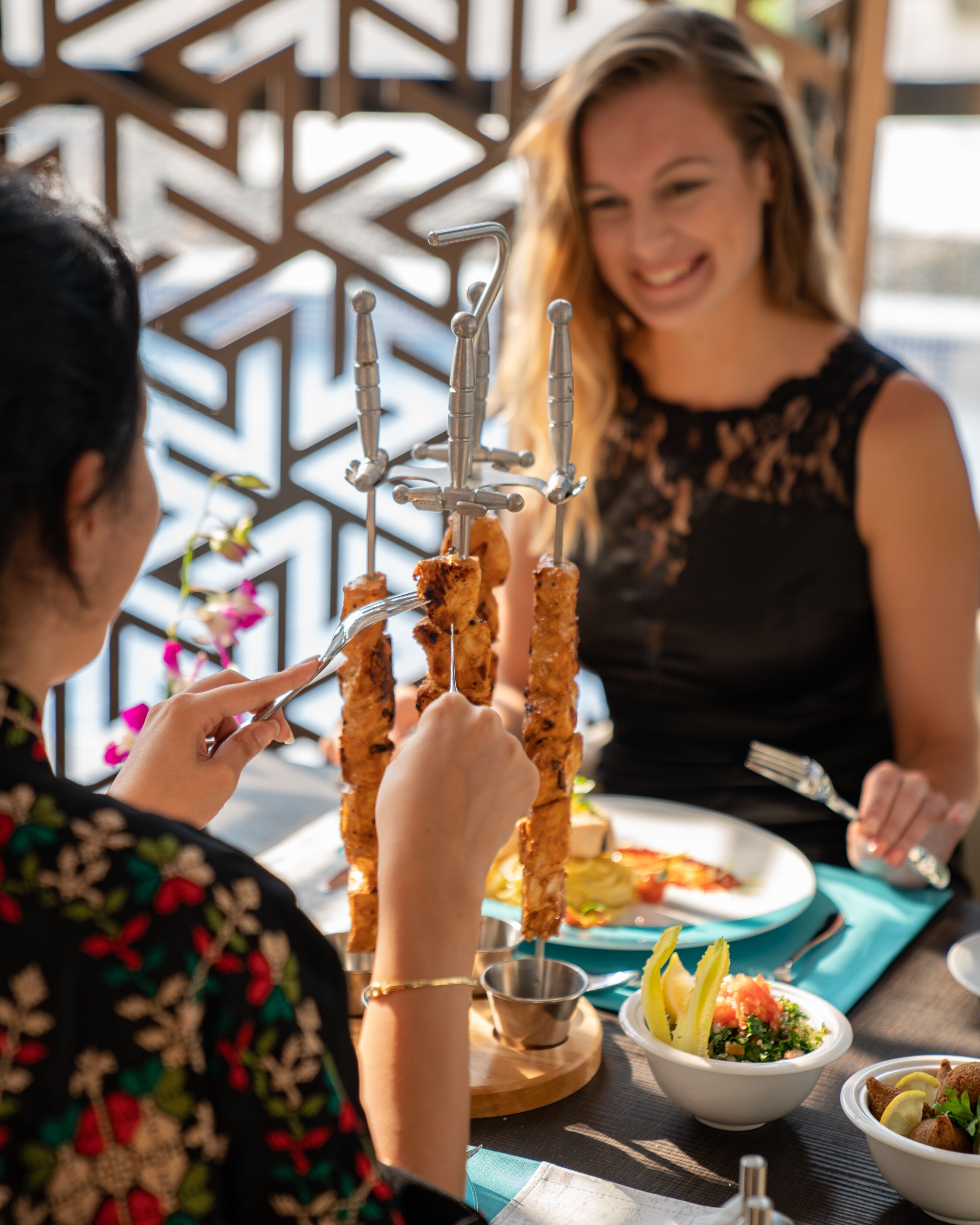 Un comedor con un soporte para brochetas lleno de comida, una mesa con platos y recipientes de comida, y un muro decorativo al fondo.