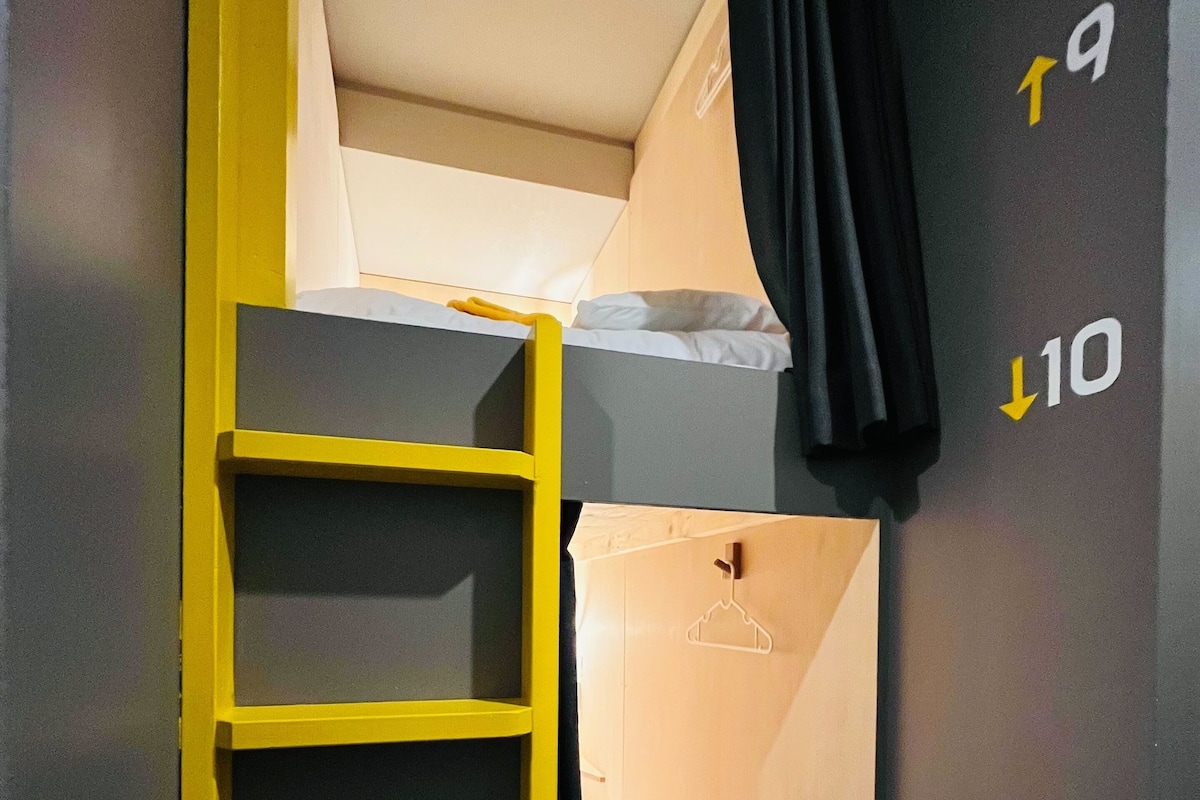 Una habitación compacta con una litera, una escalera amarilla y una cama pequeña con una almohada blanca. Se observan marcas en la pared que indican los números de habitación 19 y 10.