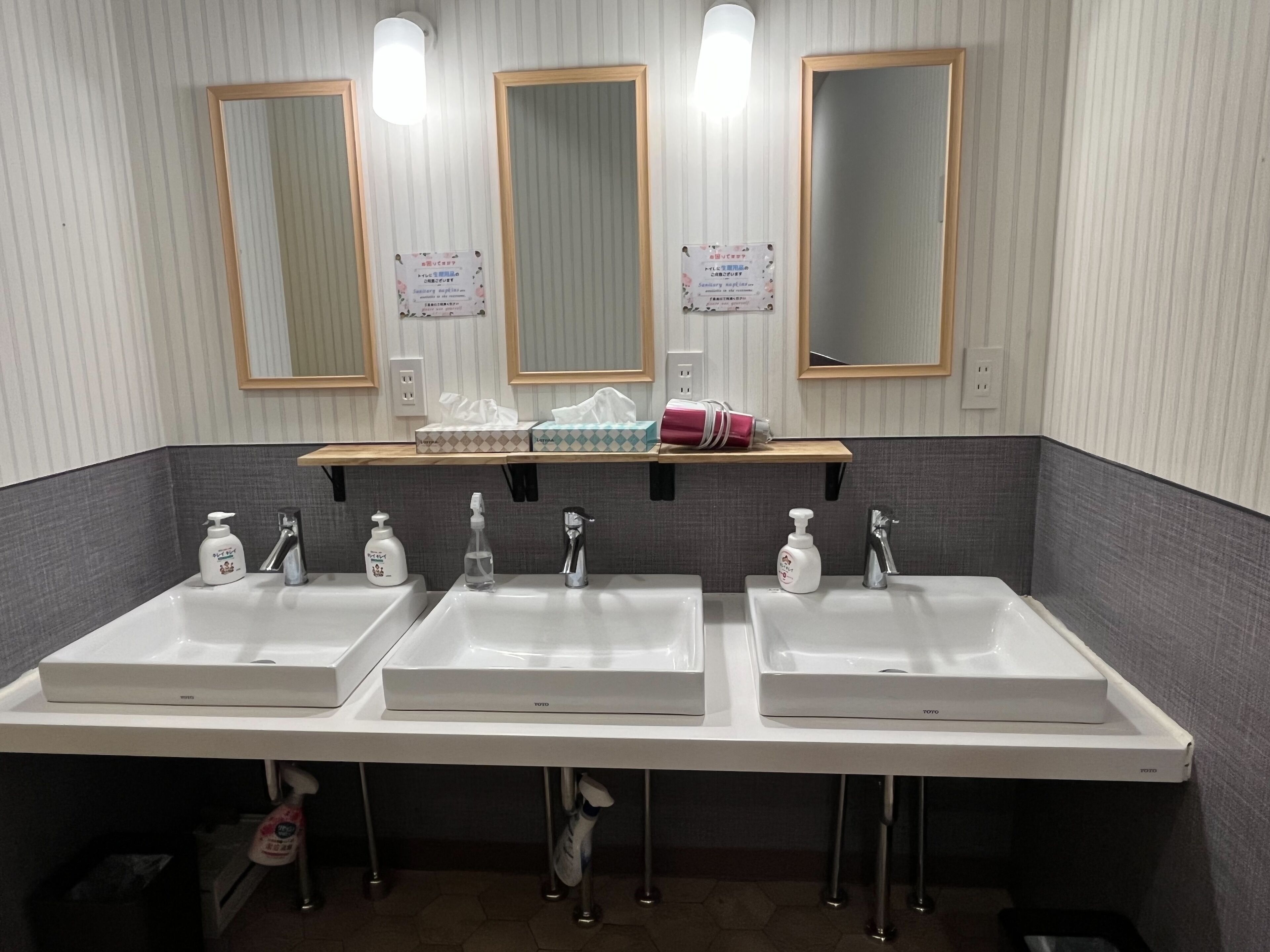 A bathroom with three sinks, each with a soap dispenser, a mirror above, and a shelf with tissues and a pink bottle.