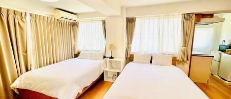 A hotel room with two beds, a desk, a chair, a lamp, a TV, and a small kitchenette.