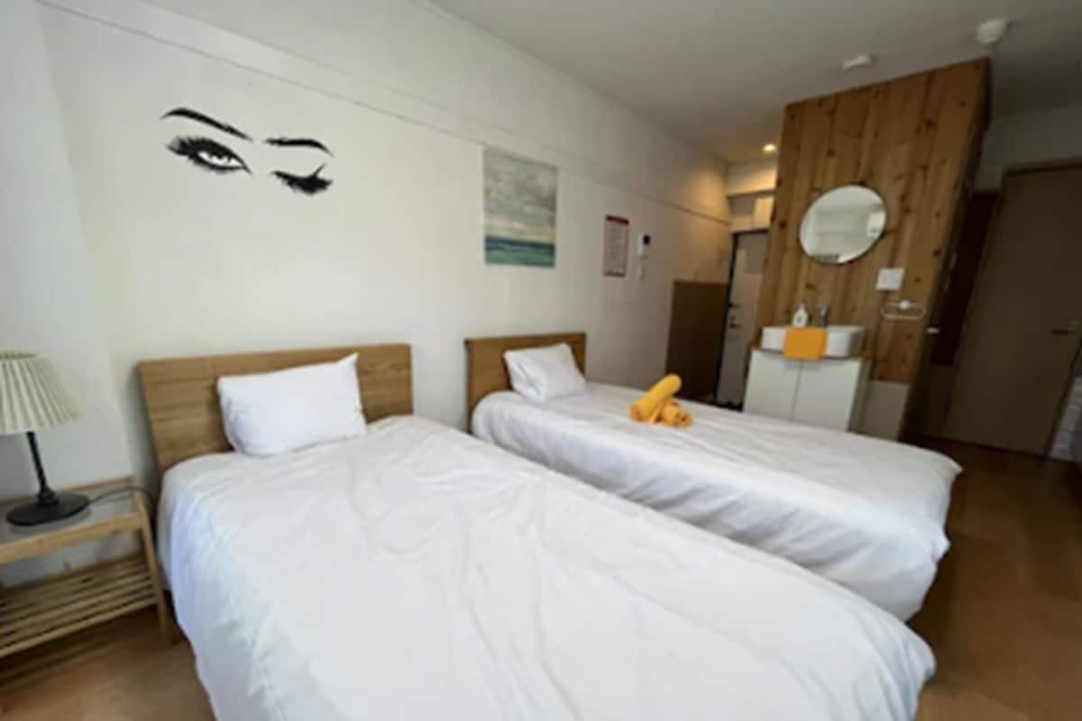 Habitación de hotel con dos camas, cabecera de madera, mesita de noche con lámpara, espejo y un lavamanos pequeño.
