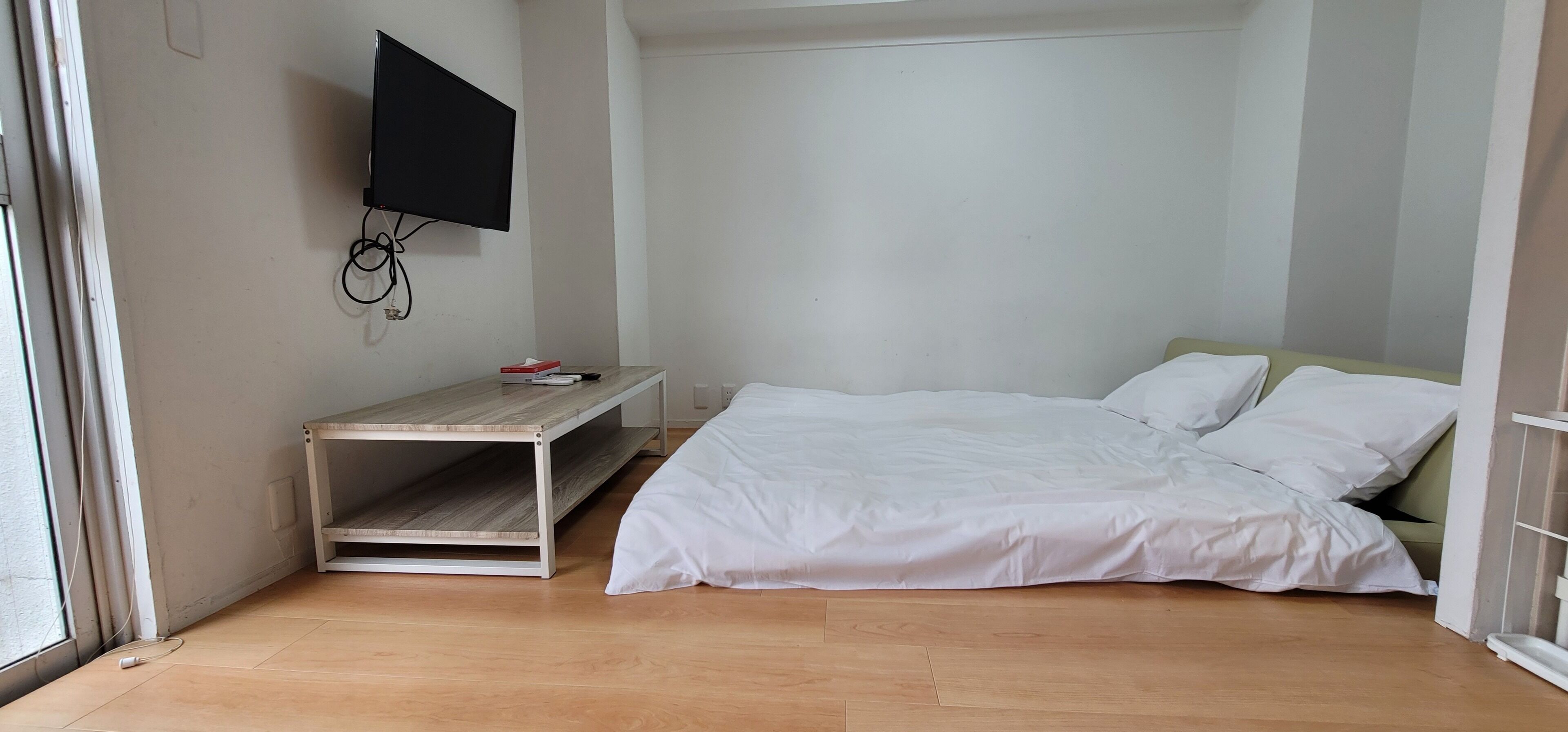 A bedroom with a bed, a TV mounted on the wall, a small table, and a shelf unit.
