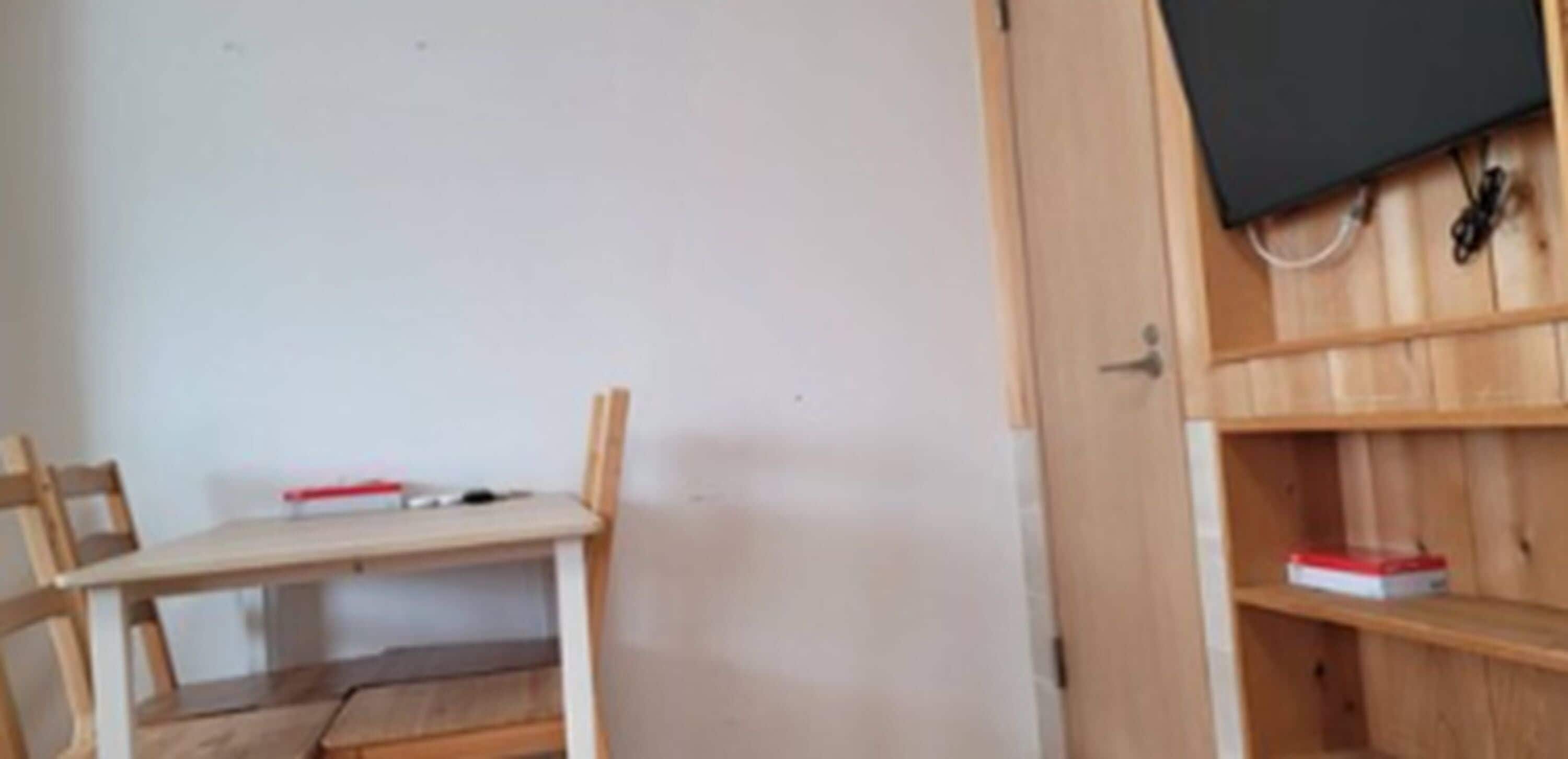 A room with a wooden desk, a chair, a television mounted on the wall, and a shelf with a red box.